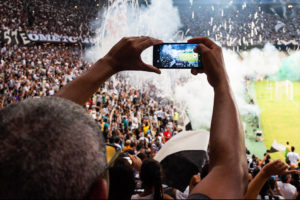 Man recording a live football game while smoke from fireworks fills the stadium.